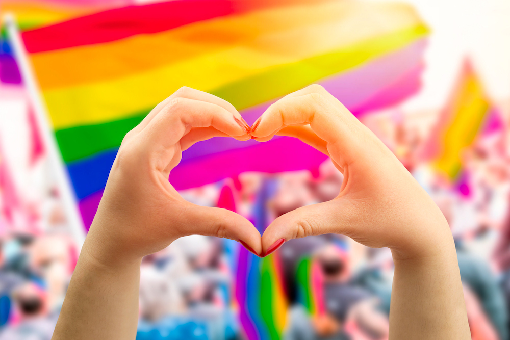 person making heart shape with hands with a rainbow Pride flag in the background